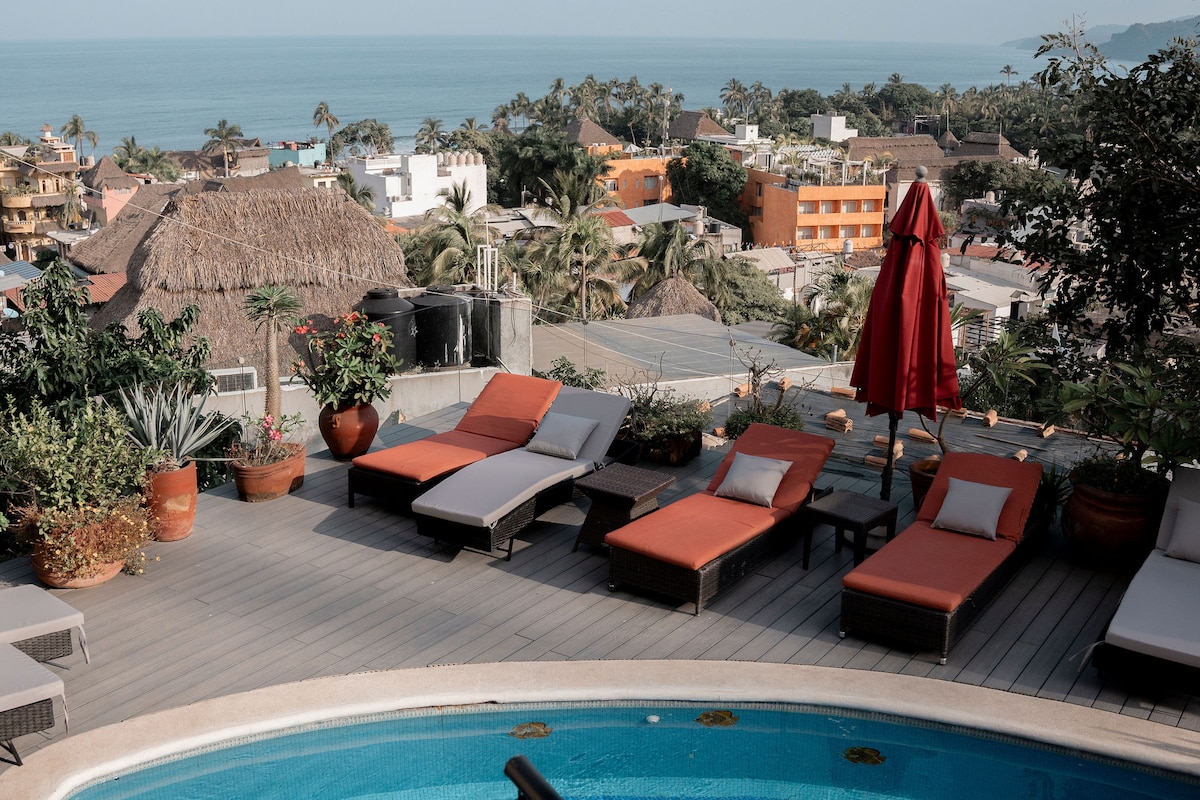 A terrace area showcases several lounge chairs, some in orange and others in gray, arranged around a shimmering pool. Tropical plants and terracotta pots adorn the space. A large red umbrella provides shade, with a scenic view of Sayulita's coastline and rooftops in the background.