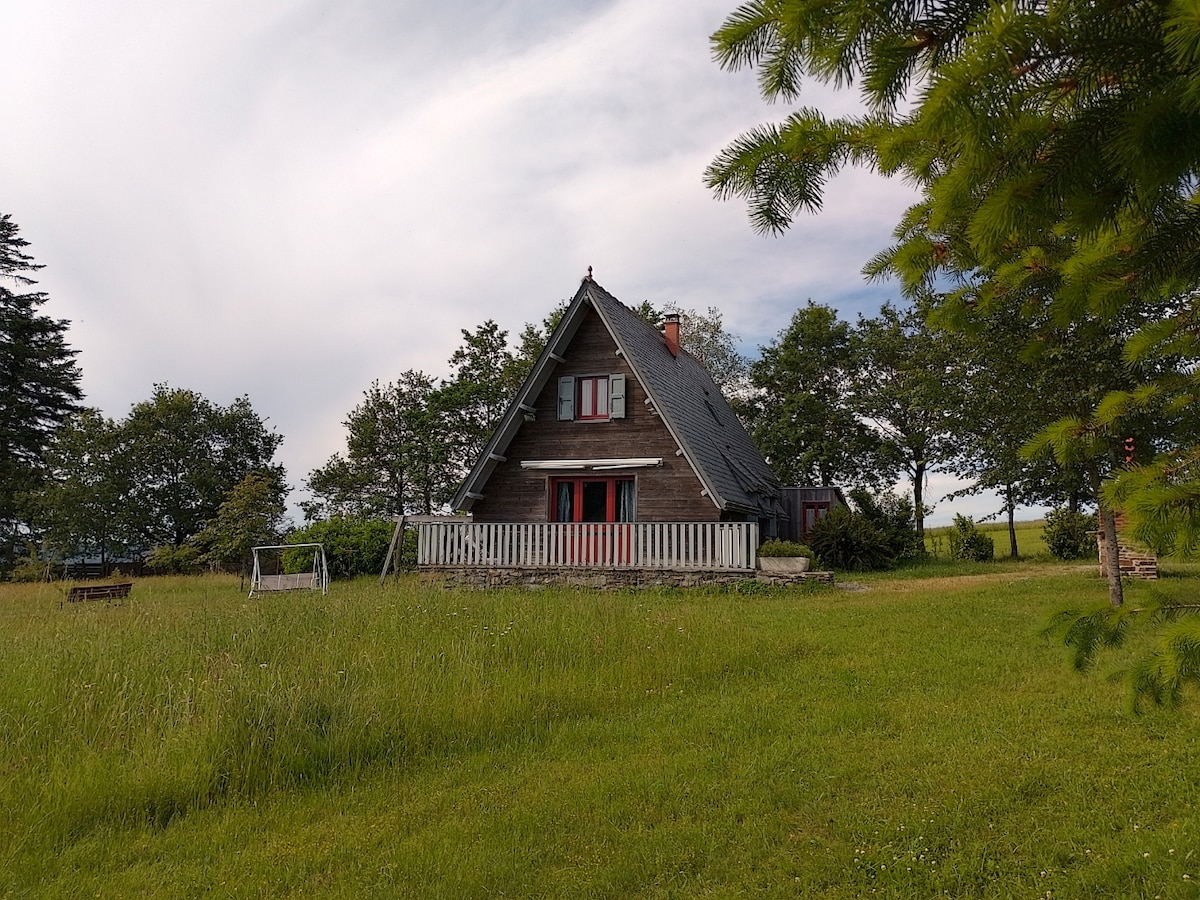 A quaint chalet is situated on a large grassy area, surrounded by trees. The structure features a steeply pitched roof and a welcoming front porch with red accents. A swing set is visible in the foreground, contributing to the peaceful outdoor environment.