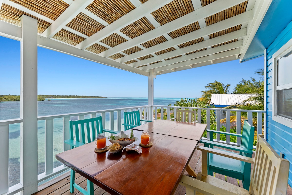 A spacious outdoor dining area is featured under a slatted roof, overlooking serene water views. A wooden table is set with drinks and light snacks, surrounded by colorful chairs. Lush greenery and distant palm trees frame the scene, enhancing the tranquil atmosphere.