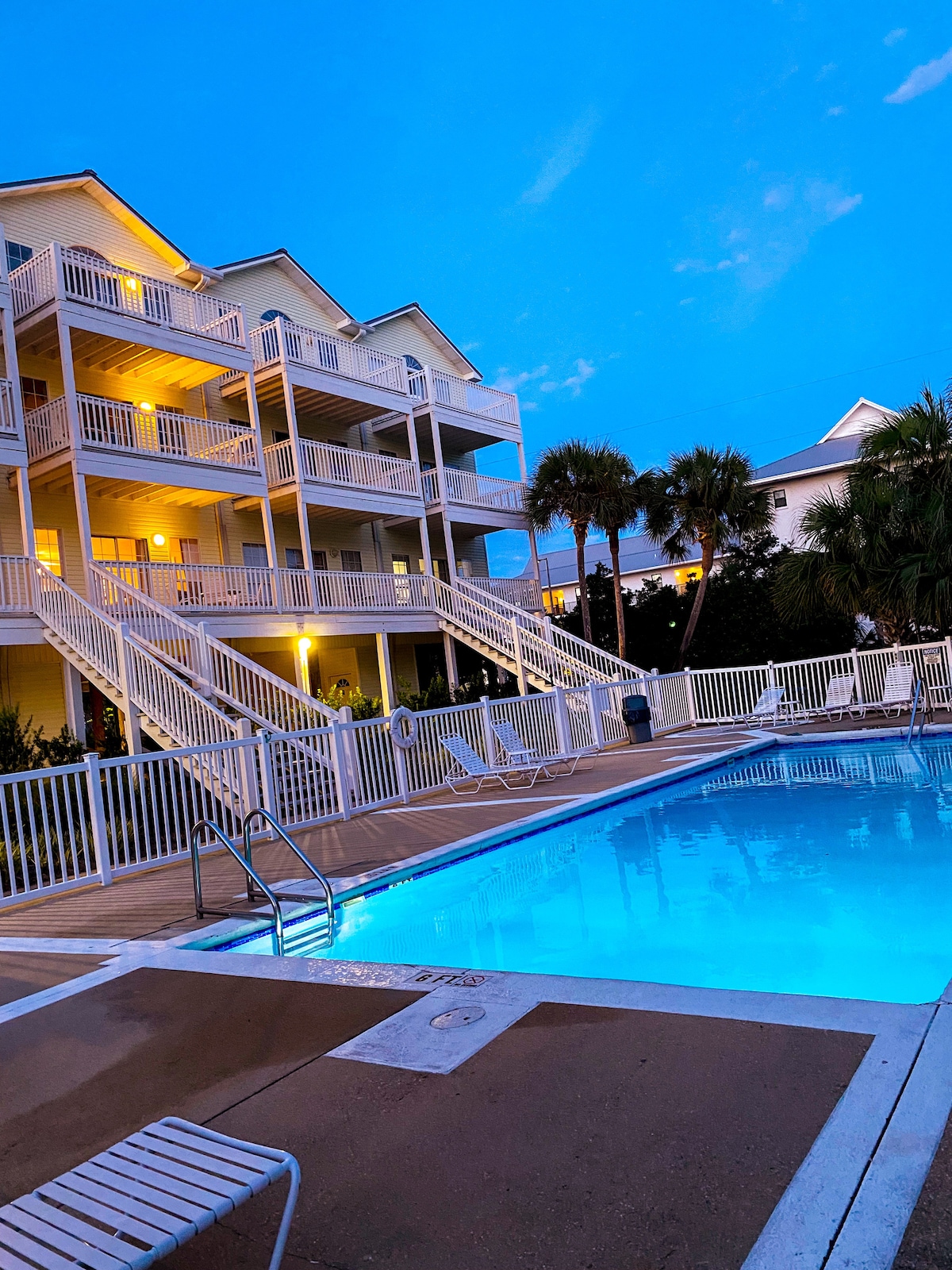 A well-lit pool area is situated in front of a three-story beach house, featuring white railings and balconies. The inviting swimming pool reflects blue hues under a twilight sky, while palm trees frame the scene, enhancing the relaxed ambiance of the outdoor space.