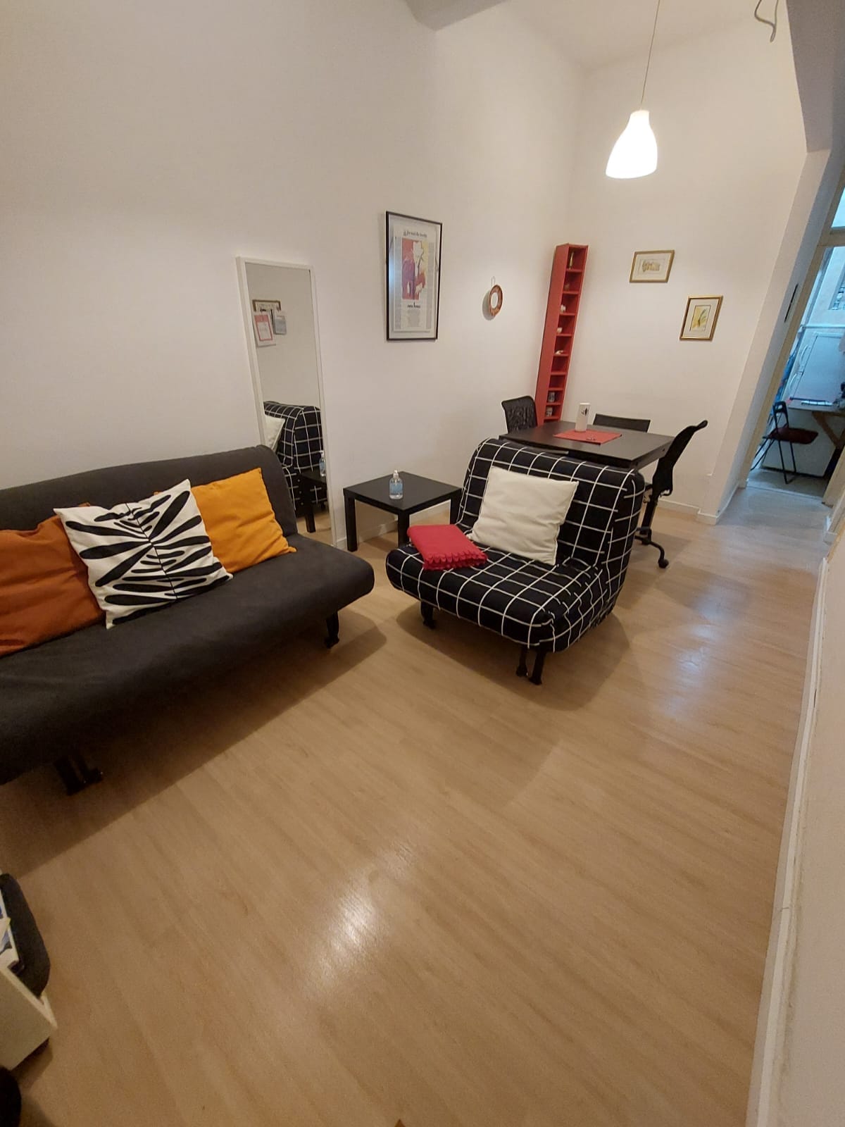 The living area includes a black futon sofa adorned with colorful cushions, and a patterned armchair next to a small black table. A well-lit space features light wood flooring, a mirror on the wall, and a bookshelf displaying framed artwork.