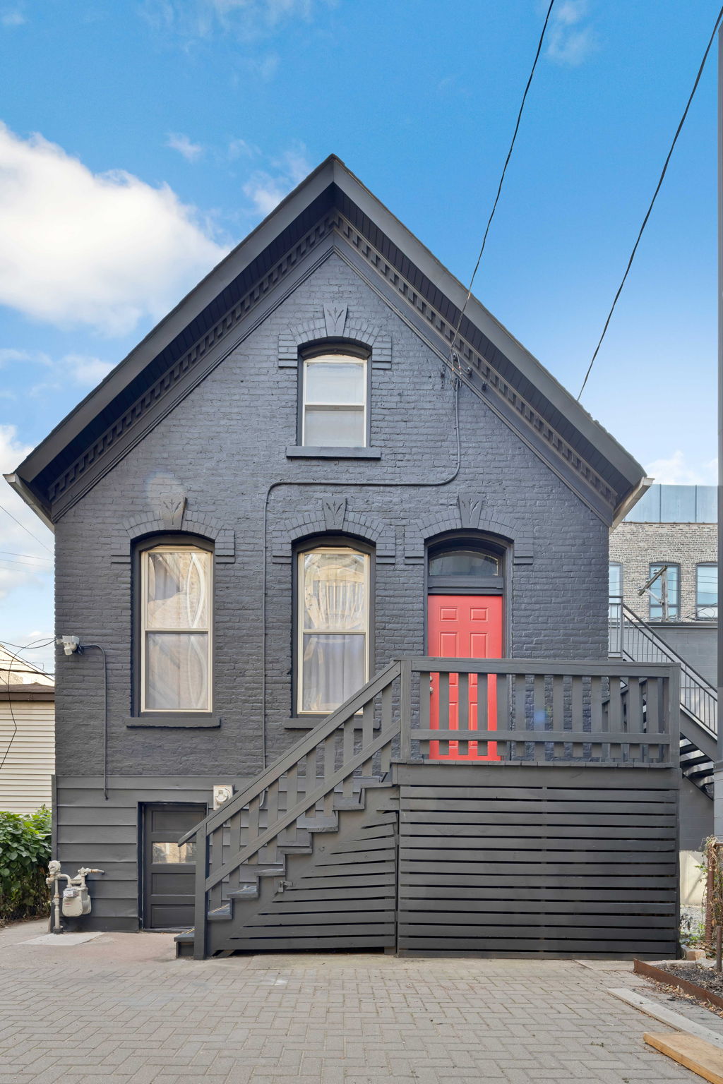 A striking, two-story brick house is presented with a dark facade and a prominent red door. Large windows provide natural light, while a set of stairs leads up to the entrance. The outdoor area features a clean pathway and surrounding minimal landscaping.
