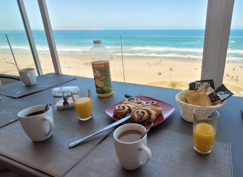 A dining table set with a view of the ocean features two cups of coffee, a glass of orange juice, and a bottle of juice. A plate holds cinnamon rolls, and a small basket contains bread. The sunlight bathes the table with a warm glow.
