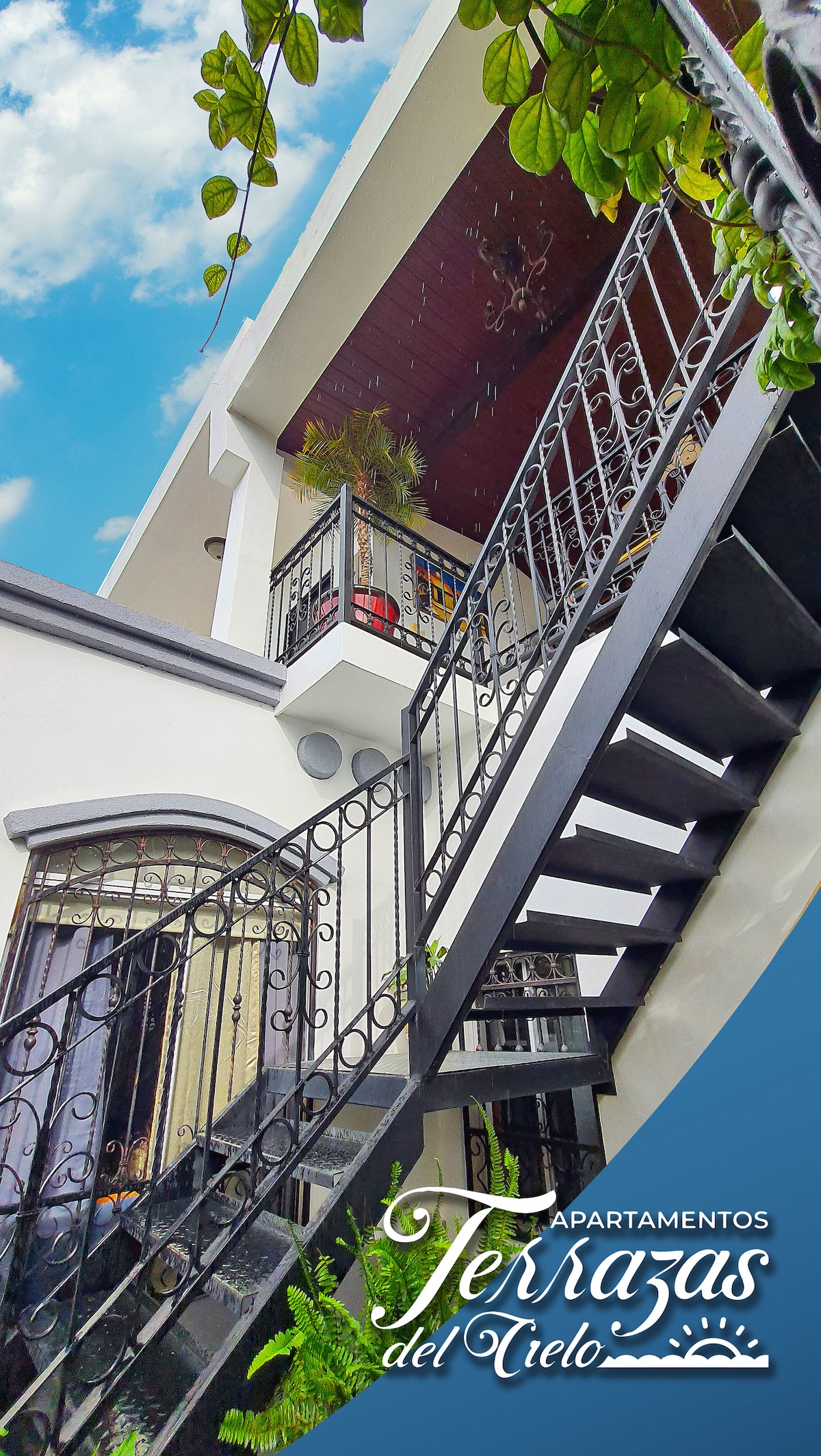 An exterior view showcases a two-story structure with a black wrought iron staircase leading to a balcony. Lush greenery lines the upper level, enhancing the building's modern architecture. Bright blue skies provide a contrast against the white walls and dark accents.
