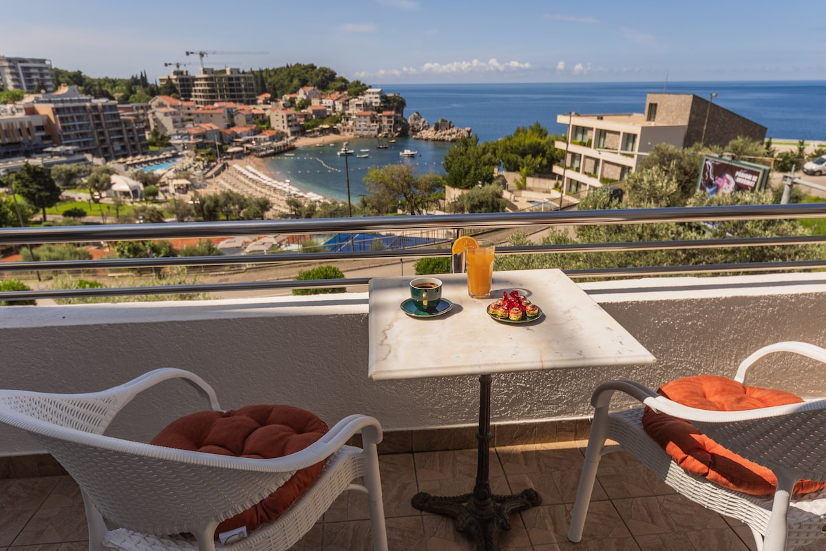 A balcony is showcased, featuring a marble table with a drink and plate of fruit. Two white lounge chairs with orange cushions provide a comfortable seating area. A scenic view of the sea and coastal town sprawls beyond, under a clear blue sky.