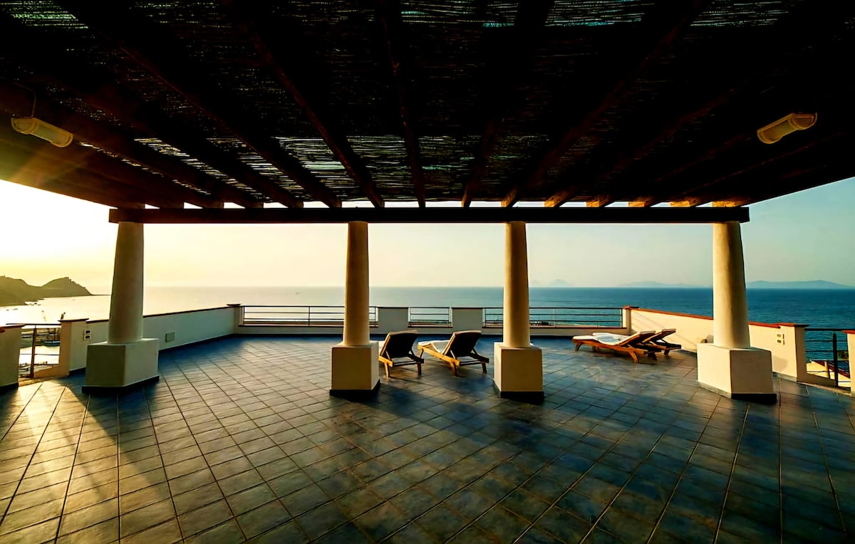 An expansive terrace is presented, featuring a shaded area under wooden beams. Sun loungers are positioned for relaxation, with panoramic views of the sea visible in the background. The surface is tiled, and the horizon showcases soft hues from the setting sun.
