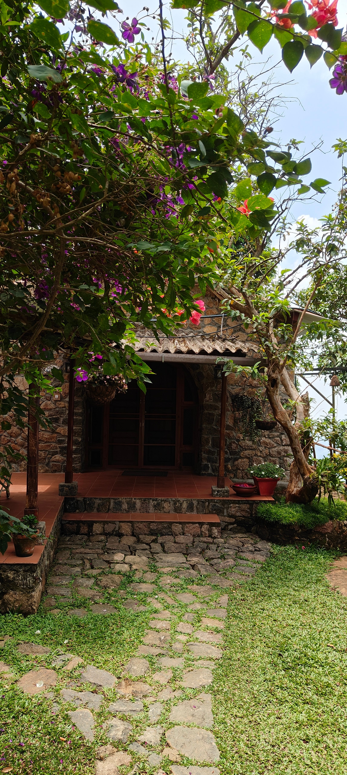 The entrance to a rustic stone cottage is framed by vibrant flowering plants and greenery. A pathway of stones leads to the inviting wooden door, while lush grass surrounds the area, enhancing the natural setting.