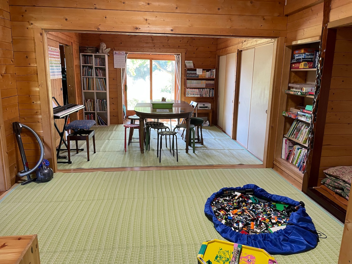 An inviting living space features wooden walls and a large window allowing natural light to fill the room. A spacious table surrounded by chairs is positioned at the center, with a colorful collection of toys on the floor and bookshelves lined with various items visible in the background.