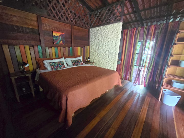 A generous king-sized bed in a room abounding in colors of the Caribe.  Headboard is crafted from a salvaged fence, original artwork by Kelly Simpson, a treasured guest.  En-suite bath through carnival curtain.