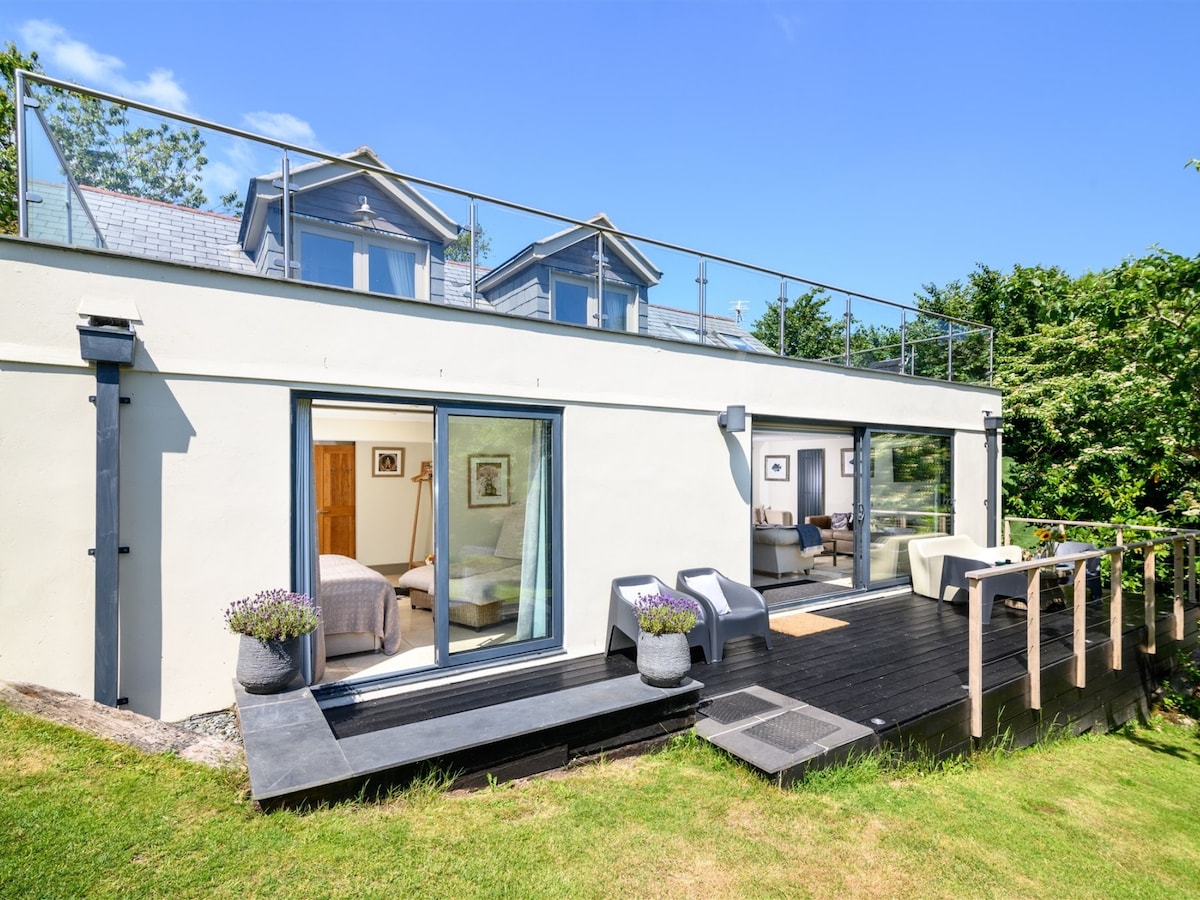 The exterior of the Summer House is shown, featuring a modern design with large glass doors that open to the deck. A railing along the upper terrace offers a glimpse of the surroundings. Flower pots are positioned on the deck, complementing the green lawn.
