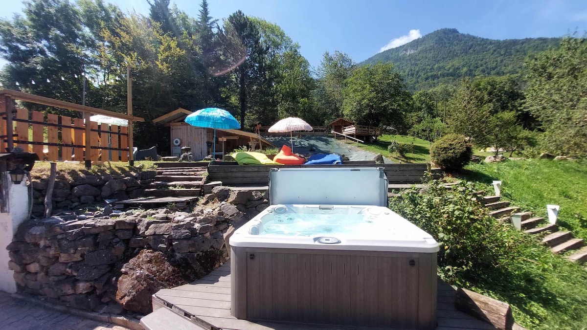 A hot tub is situated on a wooden deck, surrounded by lush greenery. Colorful sun umbrellas provide shade nearby, with lounge chairs arranged on a gently sloping area leading up to a wooded backdrop. The mountains rise in the distance under a clear blue sky.