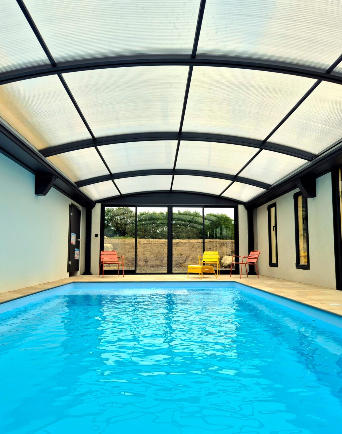 An indoor swimming pool is framed by expansive glass walls, allowing natural light to illuminate the space. Colorful chairs are positioned along one side, providing a cozy spot for relaxation. The water glistens in various shades of blue, inviting a sense of tranquility.