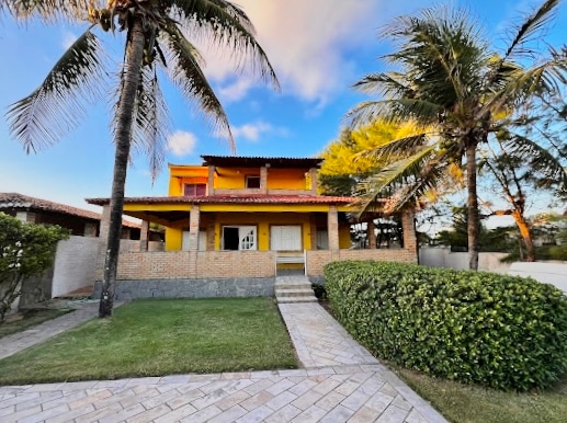 A vibrant two-story house with a yellow exterior is surrounded by lush palm trees. The front features a well-maintained lawn and a stone pathway leading to the entrance, emphasizing a welcoming atmosphere. Large windows provide ample natural light to the façade.