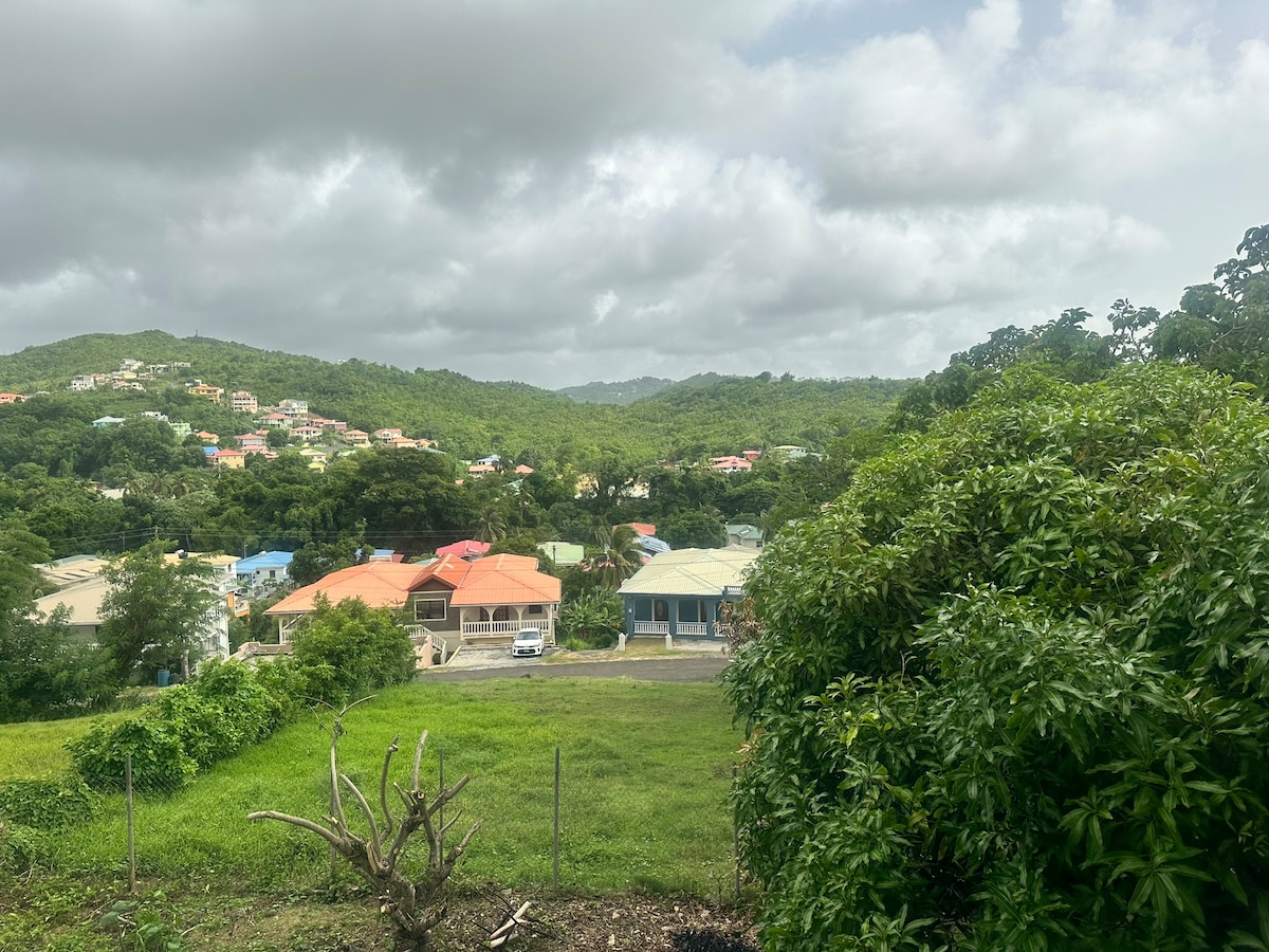 The image showcases a scenic view of a lush green landscape, featuring hills in the background and colorful rooftops dotting the neighborhood. Overcast skies provide a soft lighting effect that enhances the natural beauty of the surroundings.