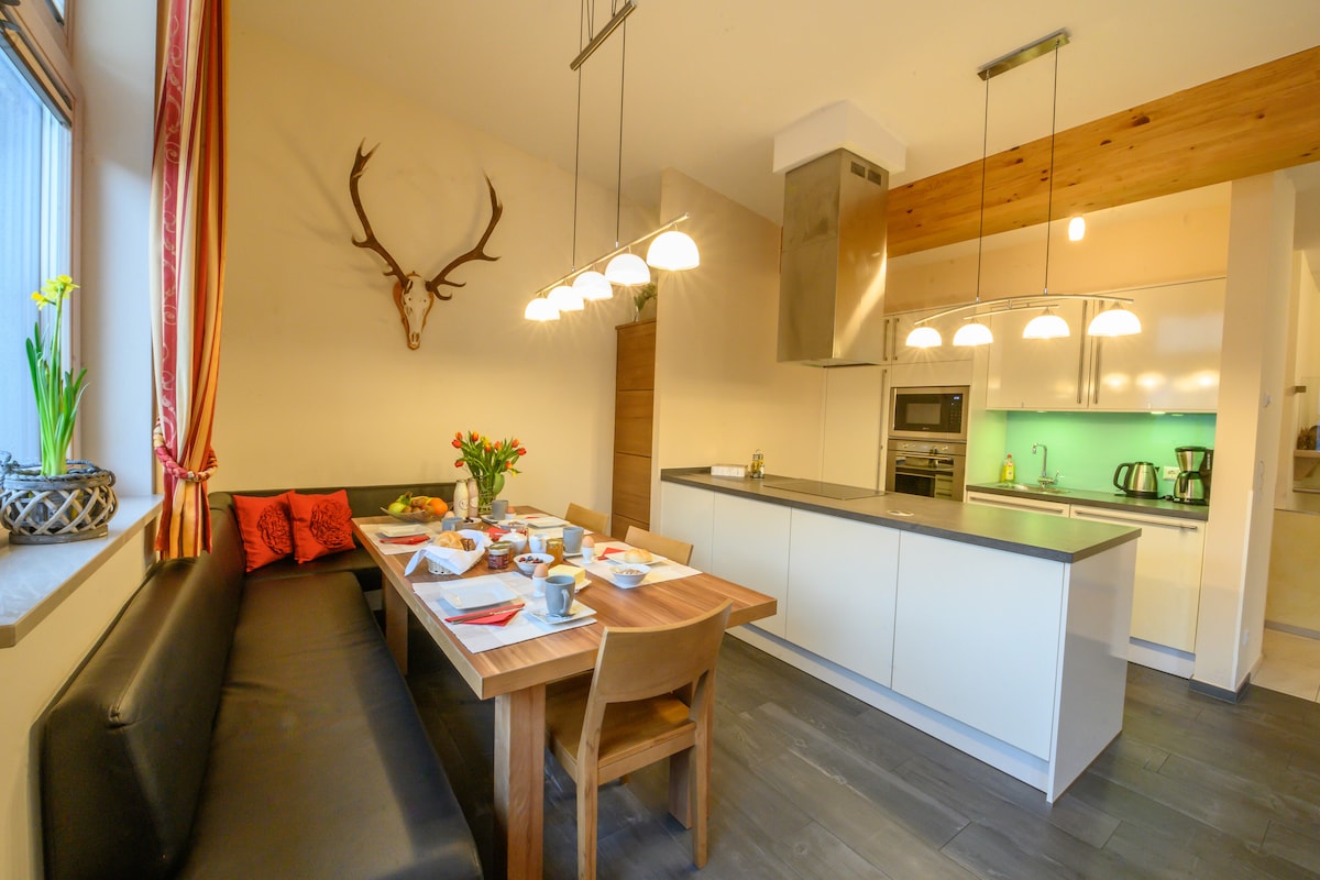 A modern kitchen and dining area feature a dark seating bench and a wooden dining table set for a meal. Overhead pendant lights illuminate the space, while a sleek kitchen counter and cooking appliances are visible in the background. Large windows allow natural light to enter.