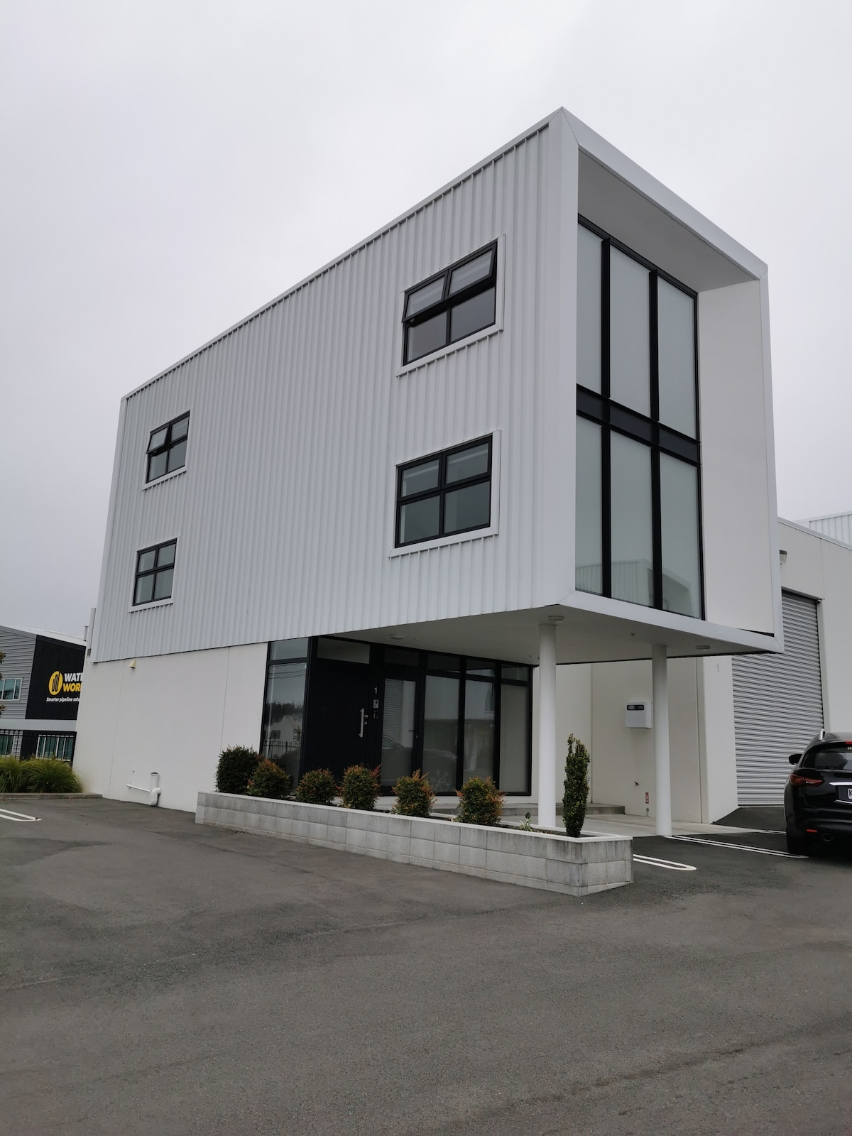 A modern, two-story building is shown, featuring a sleek white exterior with large windows. The structure is elevated on slender pillars, allowing for a clear view of the parking area. Minimal landscaping is present at the base, with neatly trimmed shrubs increasing the visual appeal.