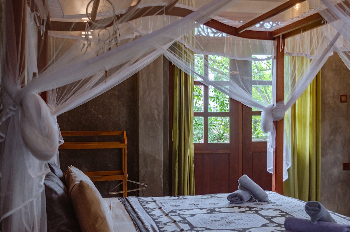 A spacious room features a king size canopy bed adorned with a mosquito net. Fresh towels are neatly placed on the bed, while natural light filters through the window, accentuating the textured walls and wooden accents.