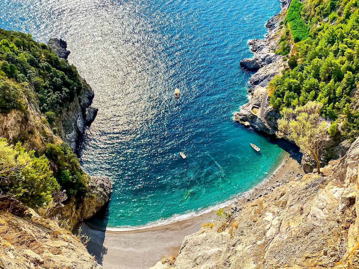 A serene cove is captured from above, featuring a sandy beach framed by rocky cliffs and lush greenery. The clear blue water gently laps at the shore, with a few boats floating gracefully on the surface.