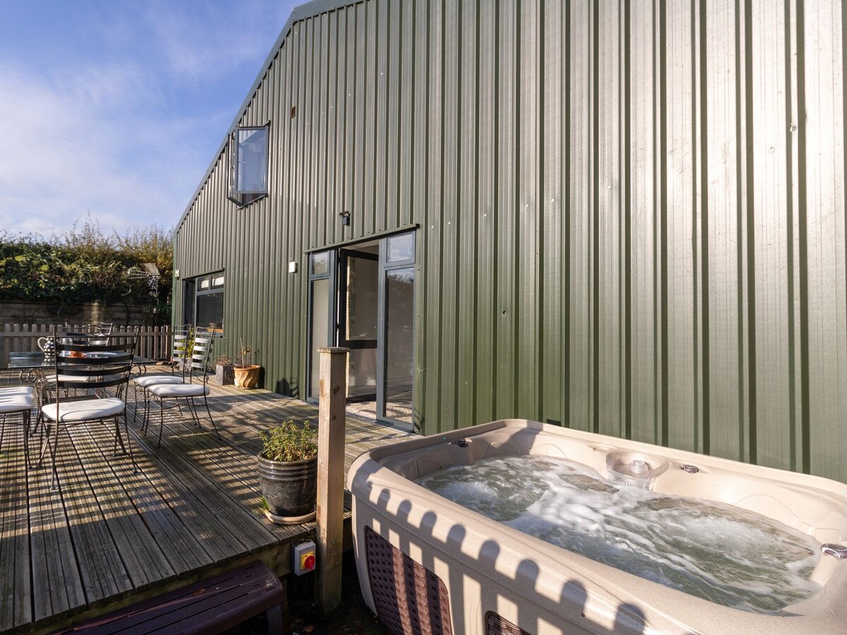 A modern hot tub bubbles invitingly on a wooden deck, alongside a round table set with six chairs. The exterior of the barn features a sleek, green metal facade with large windows reflecting natural light and the surrounding outdoor space.