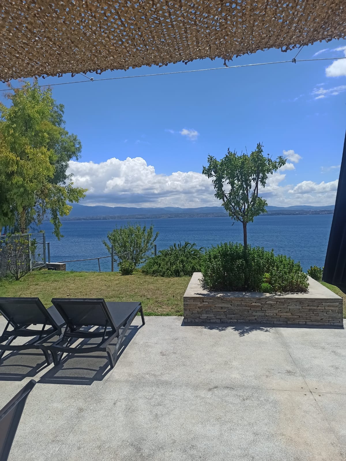 A spacious outdoor area features sun loungers positioned on a patio, overlooking the serene sea. A garden bed with various plants is present, complementing the natural landscape. The view captures a tranquil horizon with clouds reflecting the sunlight across the water.