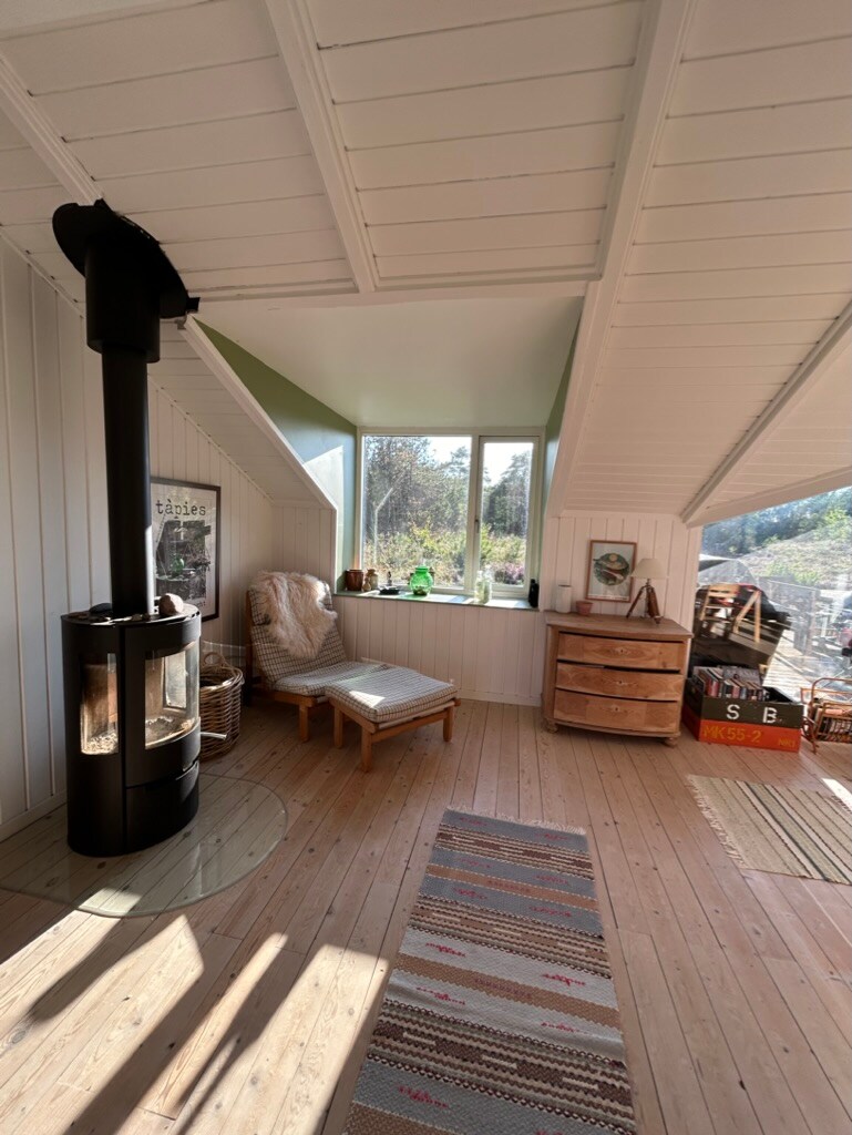A bright corner of the room features large windows allowing natural light to fill the space. A wood-burning stove is positioned near a comfortable chair and a small wooden table. A colorful area rug adds warmth to the wooden flooring.