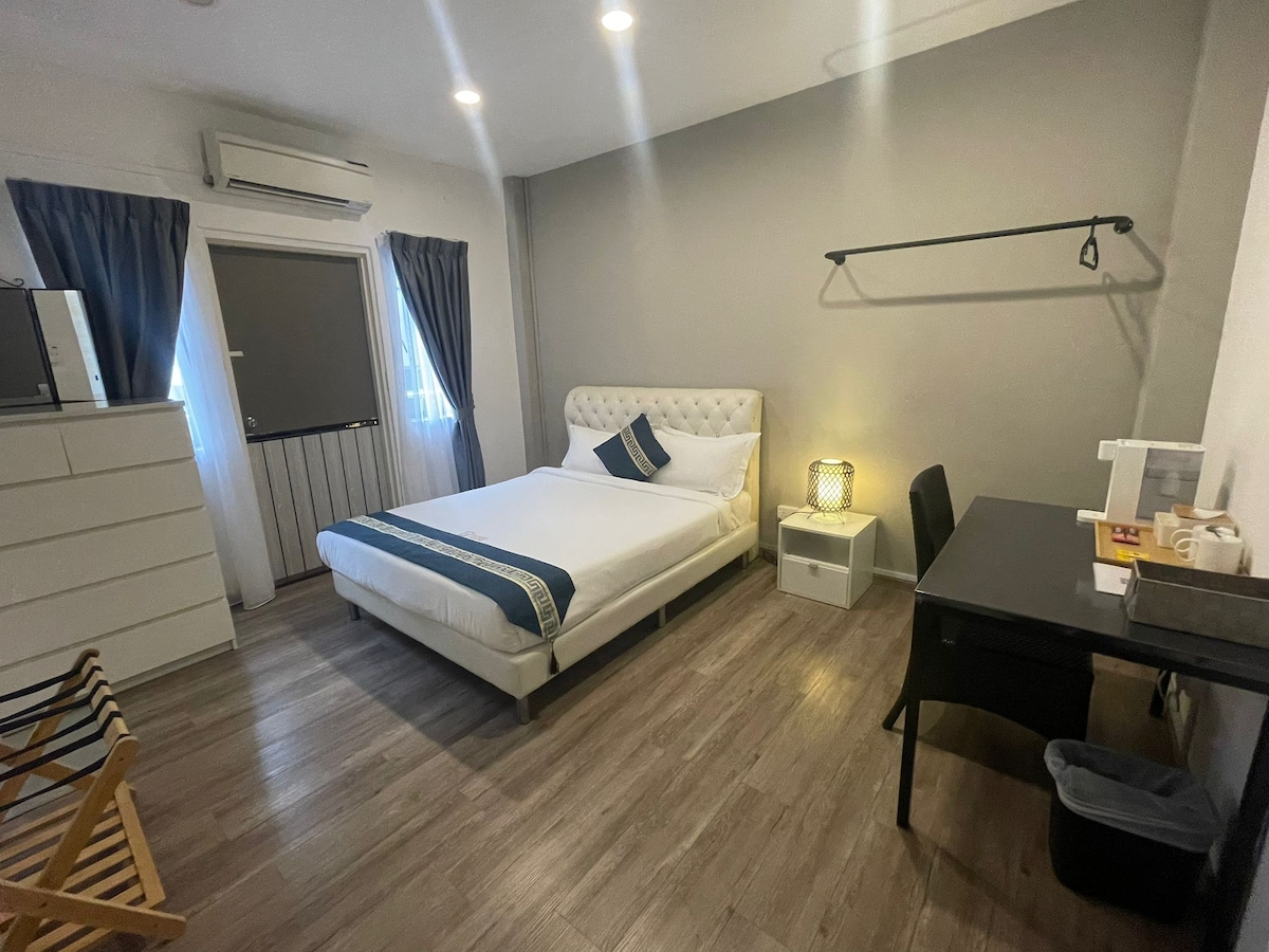A well-furnished room features a spacious bed with a blue accent stripe. A desk and chair are positioned beside a wardrobe. Natural light enters through two windows, dressed with curtains. A small light fixture offers additional illumination, creating a minimalist and functional space.