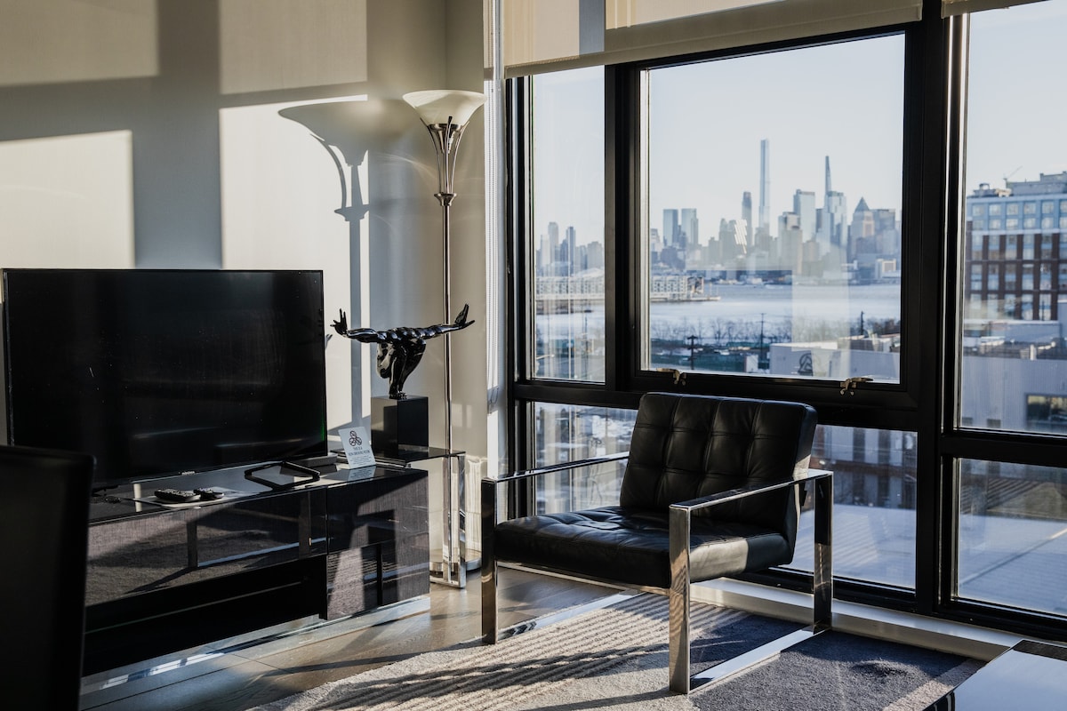 A contemporary living area features a large television mounted on a sleek console. A stylish black chair is positioned beside a large window, offering unobstructed views of the skyline and waterfront. Sunlight streams through the windows, illuminating the space.