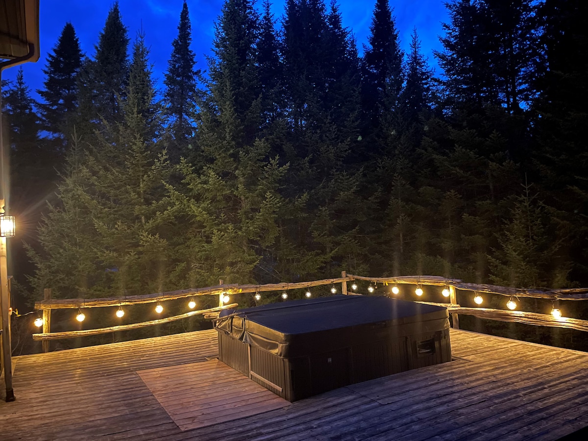 A private deck is illuminated by warm string lights, featuring a hot tub surrounded by a natural wooded landscape. Tall trees form a serene backdrop, creating a tranquil setting for relaxation under the evening sky.