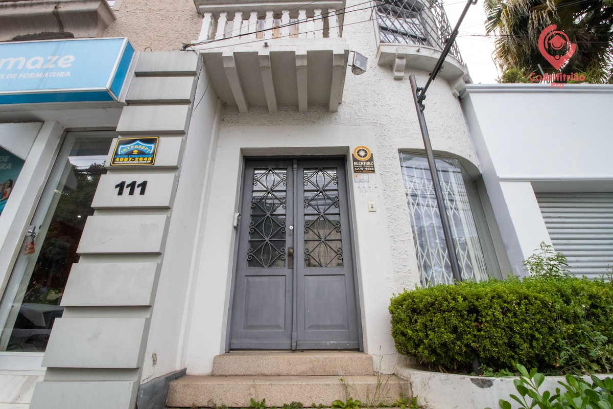 The entrance of the suite features a classic double door with decorative metal accents, leading to a cozy entryway. A small set of steps provides access, complemented by neatly trimmed greenery along the front. The facade presents a welcoming appearance in a well-maintained neighborhood.