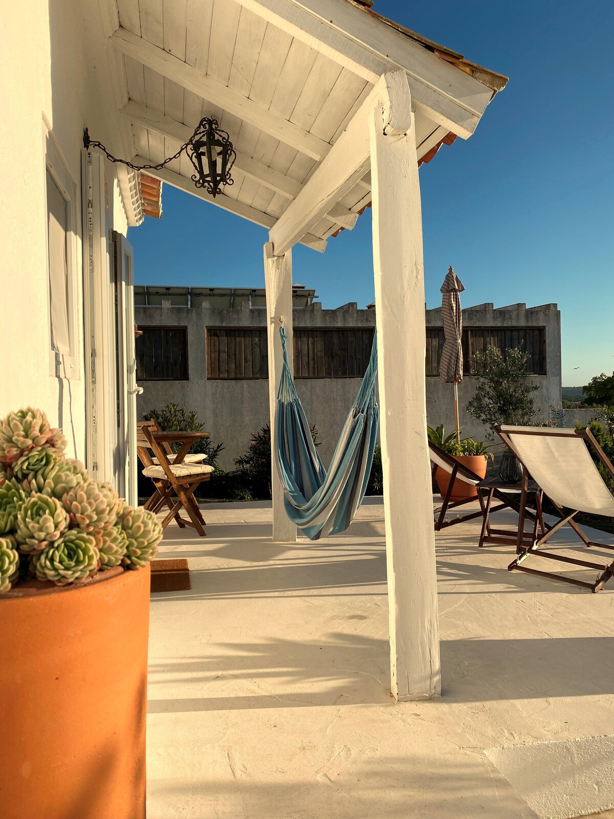 A covered outdoor terrace is presented with a blue hammock and two lounge chairs, inviting relaxation. A wooden table and chairs are positioned nearby, surrounded by potted plants. The space is complemented by a clear sky above, enhancing the tranquil setting.