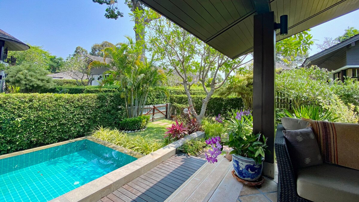 A private infinity swimming pool is surrounded by lush greenery, with tropical plants and trees framing the space. Comfortable seating is visible near the pool, providing a serene area for relaxation. The clear water reflects the vibrant colors of the surrounding foliage.