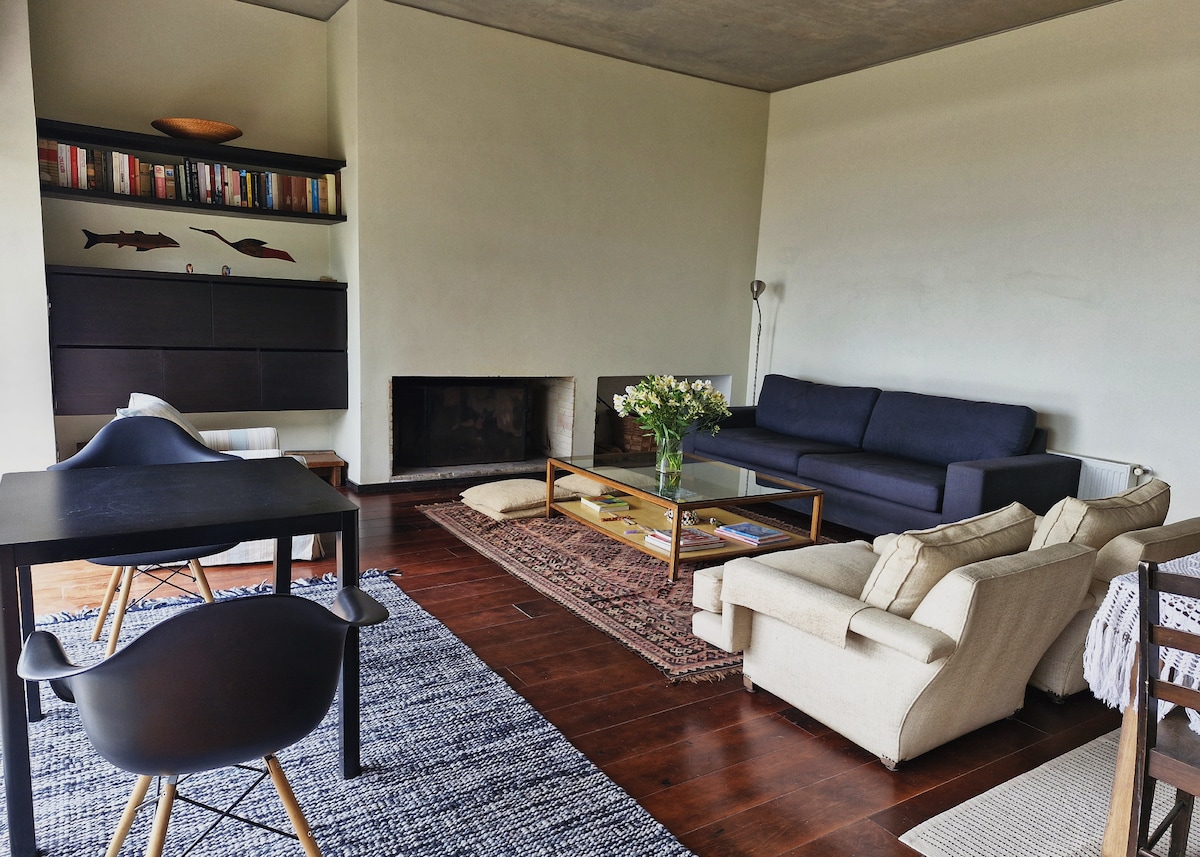 A spacious living area features a dark blue sofa and an armchair, complemented by a wooden coffee table. A rug lies on polished wooden floors, and a fireplace is positioned against the wall. Bookshelves display various books in the background, adding to the room's character.