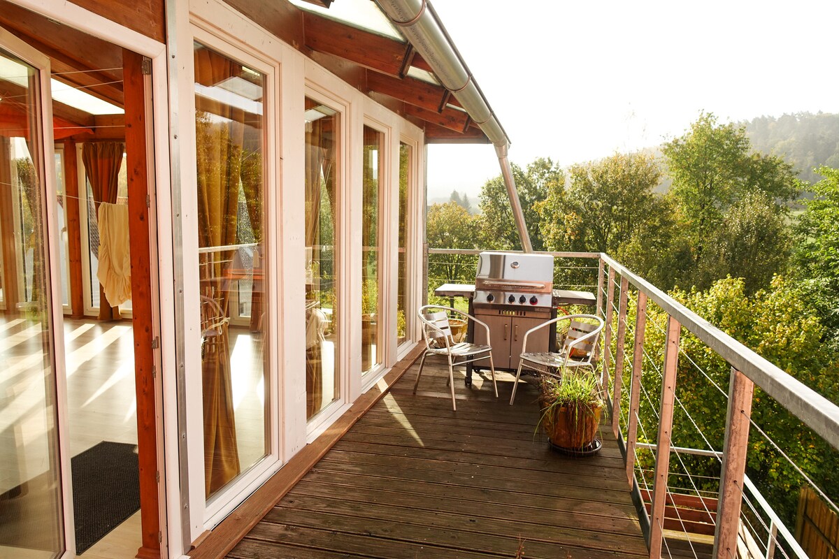 A spacious balcony features a grill and seating area, overlooking lush greenery. Large glass doors provide access to the interior, allowing natural light to fill the space. The balcony's wooden flooring complements its inviting atmosphere.