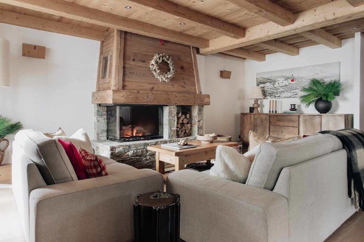 A spacious living area features a stone fireplace surrounded by wooden beams. Two comfortable sofas are positioned invitingly, with a table for gatherings. Natural elements, such as a wreath and decorative greenery, complement the warm ambiance of the room.