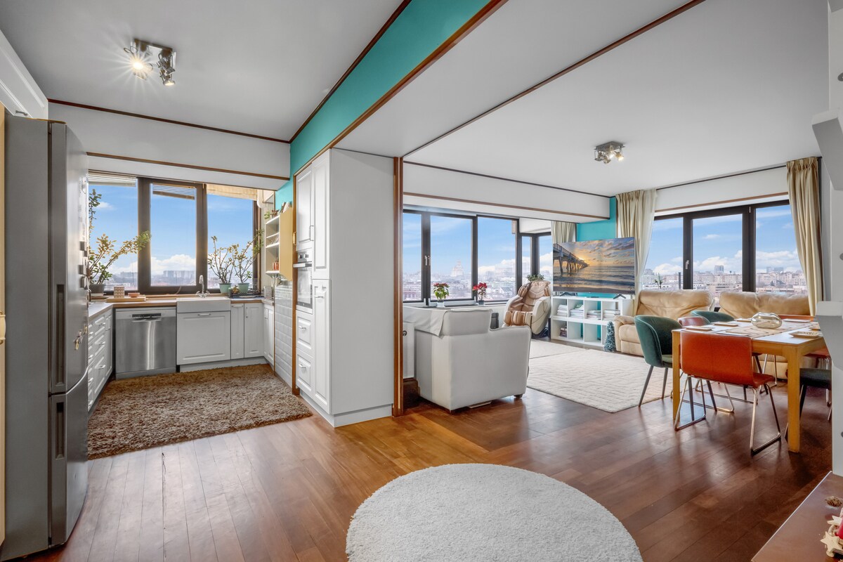 A bright and spacious open-plan living area is visible, featuring large windows that offer unobstructed city views. The modern kitchen includes stainless steel appliances, while the cozy seating area is complemented by natural light and elegant decor. A dining table with colorful chairs is also present.