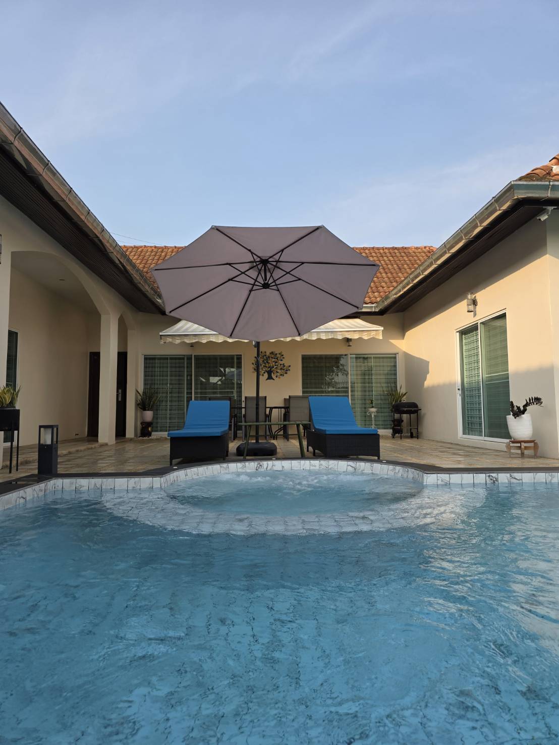 A serene outdoor space features a refreshing pool with a circular spa. Two lounge chairs are positioned under a large umbrella, with a small table nearby. The area is surrounded by modern architecture and large glass windows, creating a relaxed setting for gatherings.