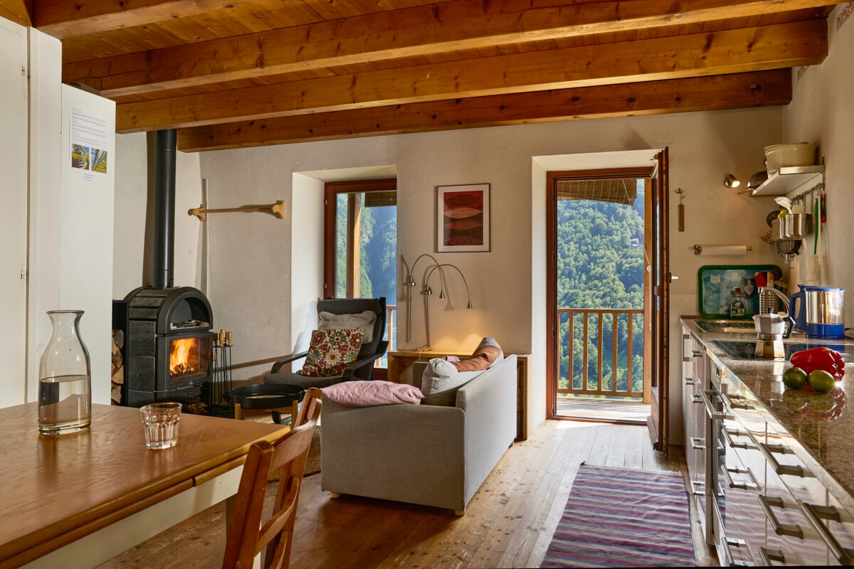 An inviting kitchen and living area features wooden beams overhead and a cozy wood stove. A dining table is positioned near the window, while comfortable seating accompanies a warm ambiance. Natural light streams in through open doors, showcasing scenic mountain views and a welcoming atmosphere.