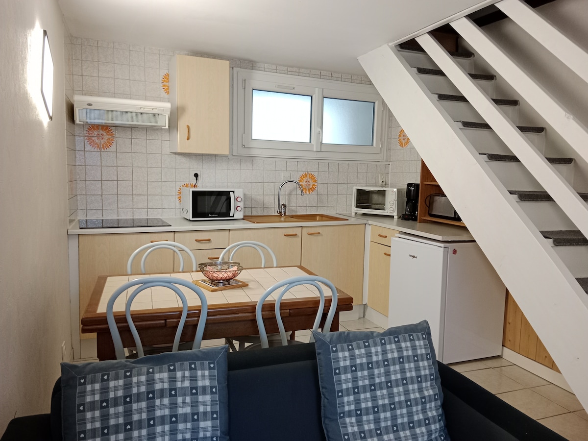 A functional kitchen area is visible, featuring light wooden cabinetry and a dining table with four chairs. Essential appliances, including a microwave and refrigerator, are present. Natural light enters through two windows, creating a bright atmosphere. A staircase is positioned in the background.