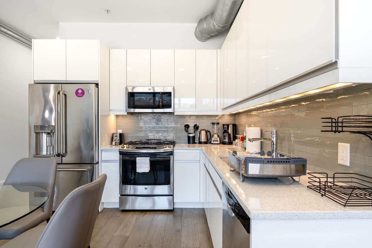 A modern kitchen is equipped with sleek white cabinetry and stainless-steel appliances, including an oven, microwave, and refrigerator. A polished countertop displays a coffee maker and utensils. Adjacent, a dining area features a glass table surrounded by stylish gray chairs, complementing the contemporary design.