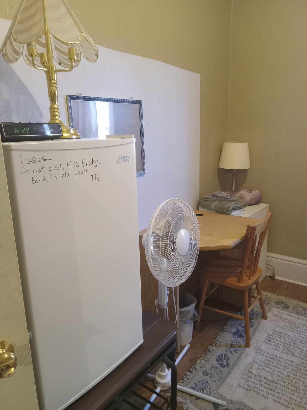 A corner of a guest room is shown, featuring a white mini fridge beside a small wooden table and chair. A table lamp provides soft lighting, and a fan is positioned on the floor. A note on the fridge advises not to push it against the wall.