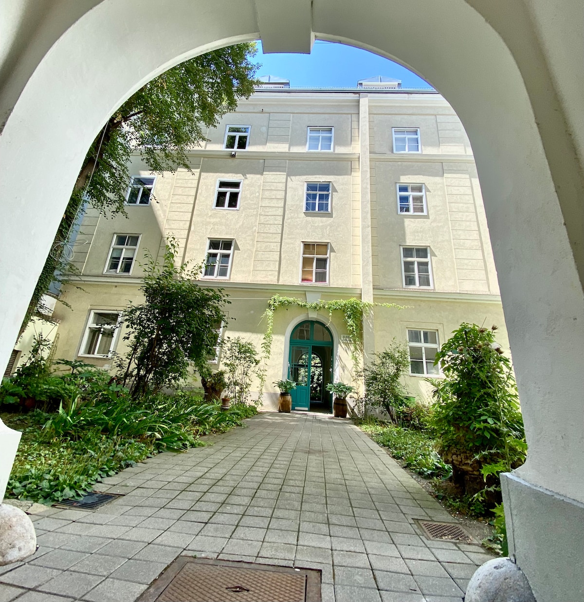 An arched entrance leads to a serene courtyard featuring vibrant greenery and potted plants. The beige-walled building displays multiple windows, allowing natural light to enhance the tranquil atmosphere. The pathway, bordered by lush plants, guides visitors through the welcoming outdoor space.