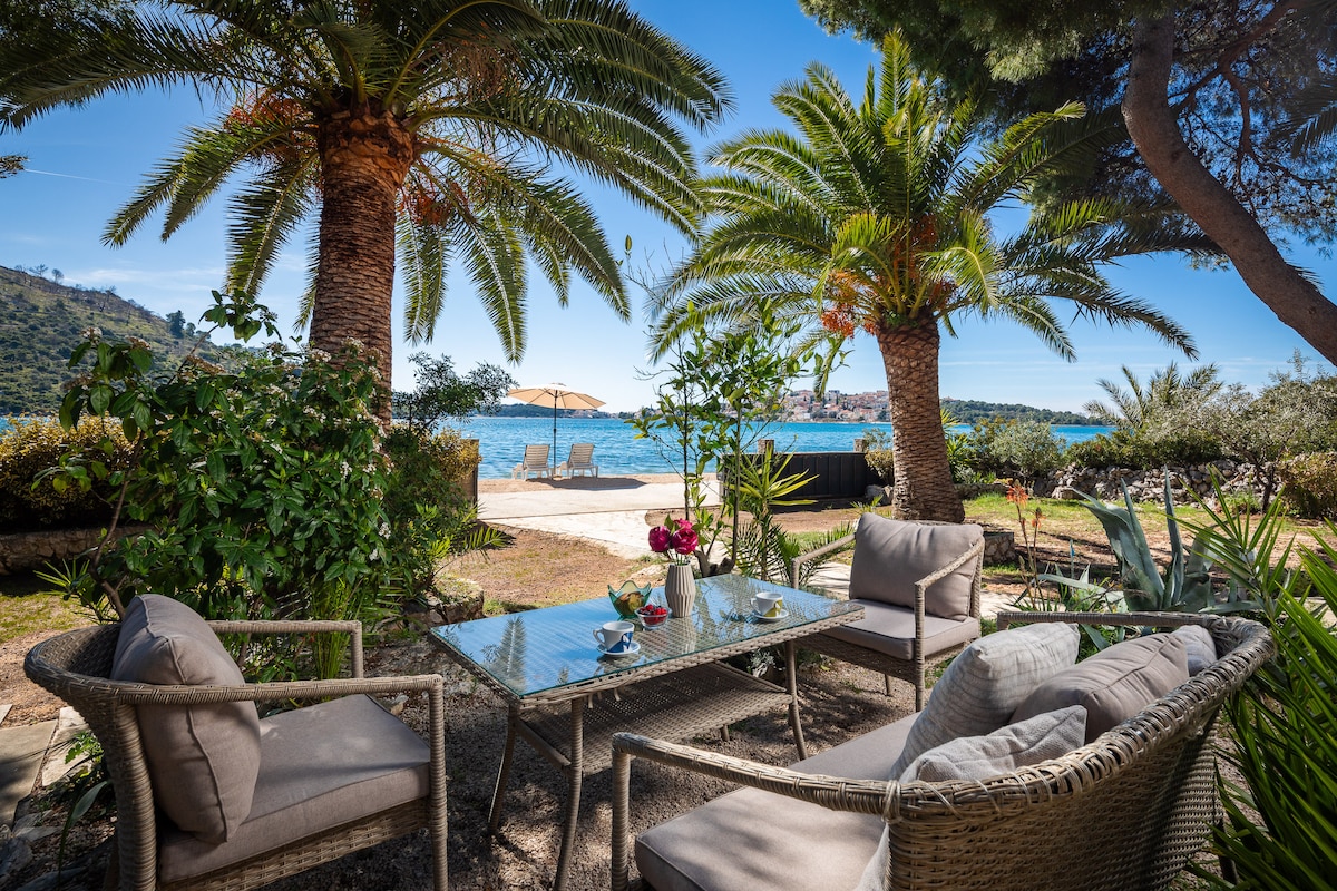 A serene outdoor seating area features comfortable wicker chairs and a glass table, surrounded by lush greenery and palm trees. An umbrella and sun loungers are visible in the background, with views of the tranquil Adriatic Sea beyond.