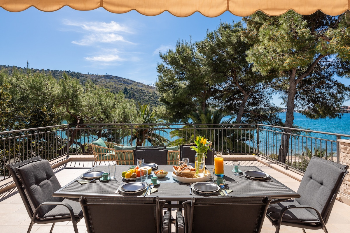A spacious terrace is set for dining with a large table and chairs, overlooking the Adriatic Sea. Plates of fresh food and drinks decorate the table, while lush greenery and palm trees frame the scenic view in the background, providing a tranquil setting.