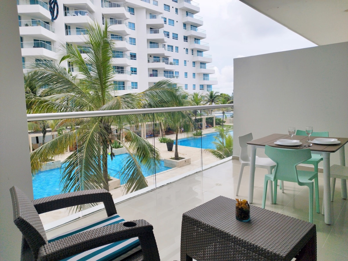 A spacious balcony features a small table with two chairs, overlooking lush palm trees and multiple swimming pools. The modern building in the background adds to the inviting environment, suitable for relaxation and enjoying the views.