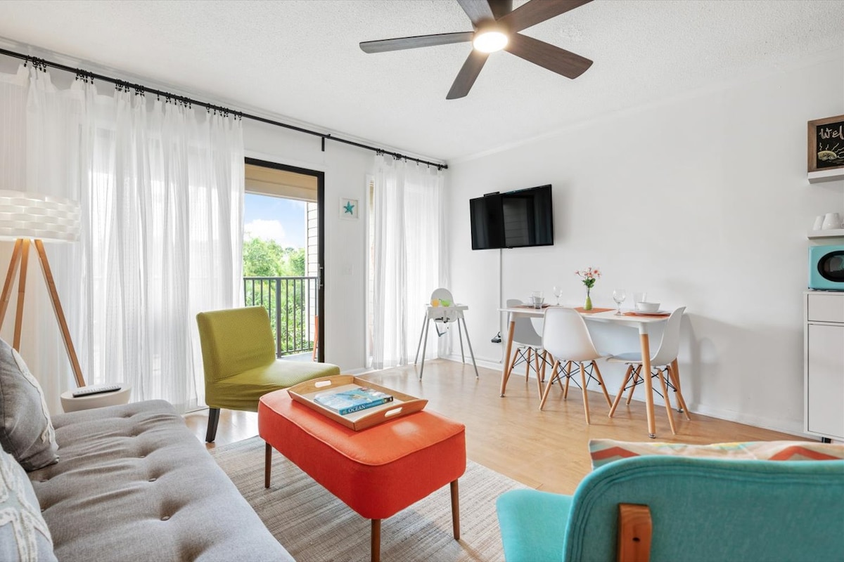 The living area features a cozy seating arrangement with a stylish sofa and colorful accent chairs. A dining table for four is present, along with a green high chair. Natural light filters through sheer curtains, enhancing the welcoming ambiance.