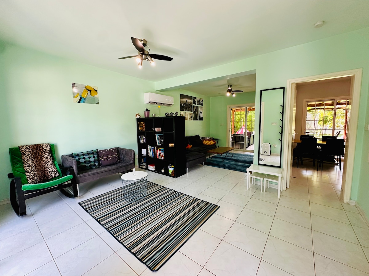 A comfortable living area features light green walls and tile flooring. A plush sofa and armchair are arranged around a central coffee table, while a bookshelf displays various items. Large windows allow natural light to illuminate the space, creating a bright and inviting atmosphere.