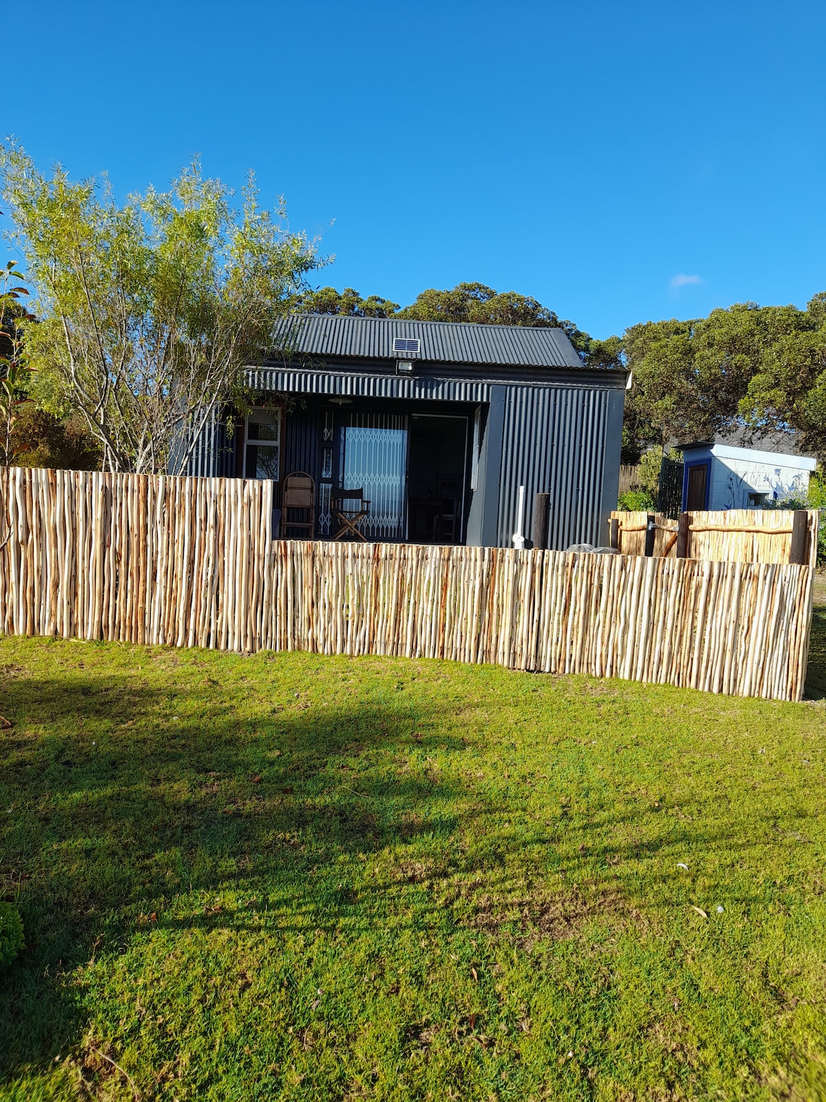 The shed-style cottage features a modern design with a dark exterior, set against a bright blue sky. A natural wooden fence surrounds the property, and a manicured lawn leads to the entrance. Large glass doors provide glimpses of the indoor space.