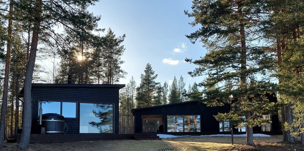A modern villa is set amidst tall trees, showcasing large windows that reflect the surrounding nature. The structure features a separate sauna building, with the calming blue sky visible in the background. The grassy area in the foreground transitions to a lightly snow-covered section.