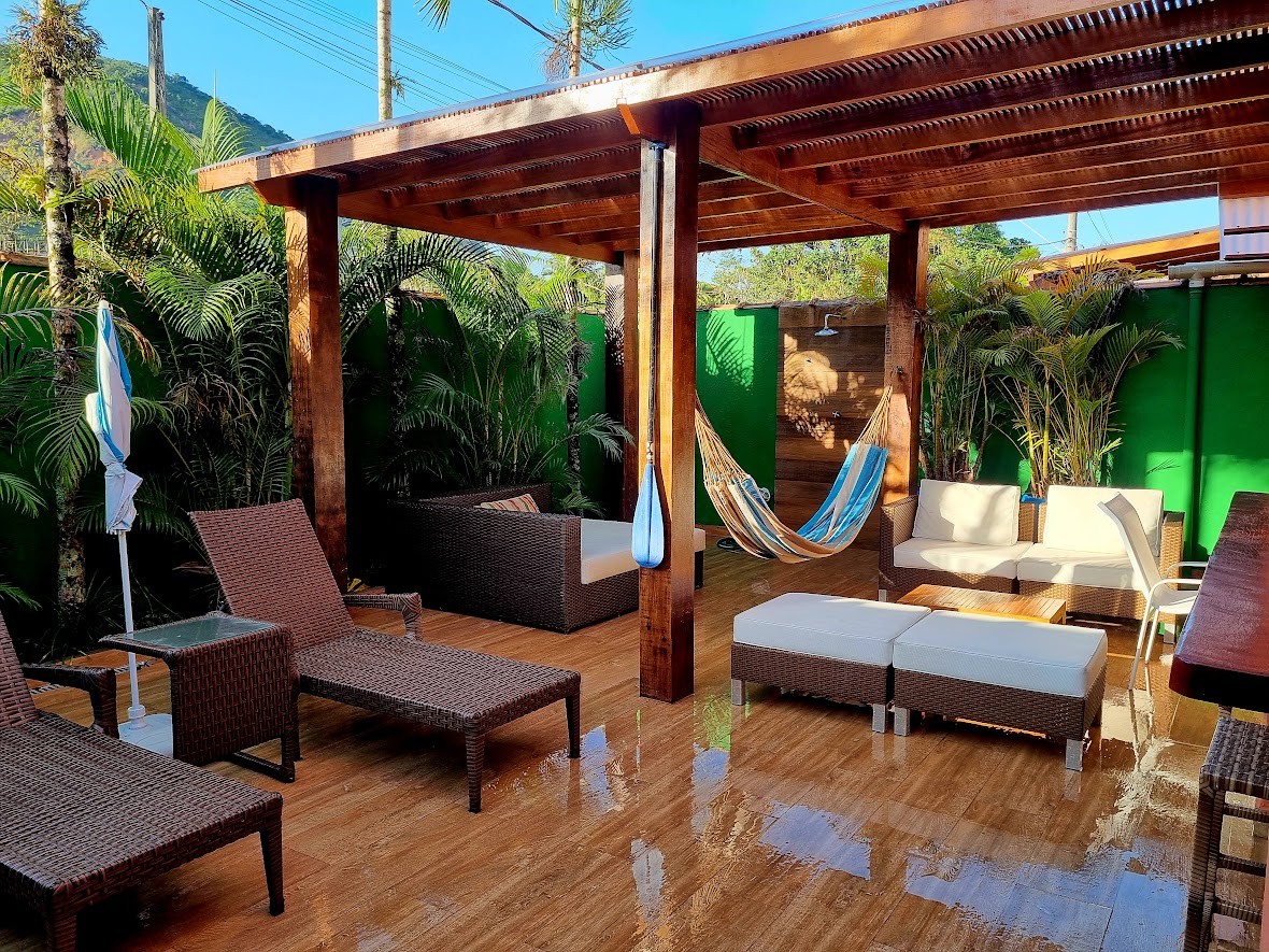 An outdoor area is highlighted by a wooden pergola, featuring a cozy lounge set with a sectional sofa and a hammock. Two sun loungers are positioned on bamboo flooring, surrounded by lush greenery, and a dining area with a table extends along one side.
