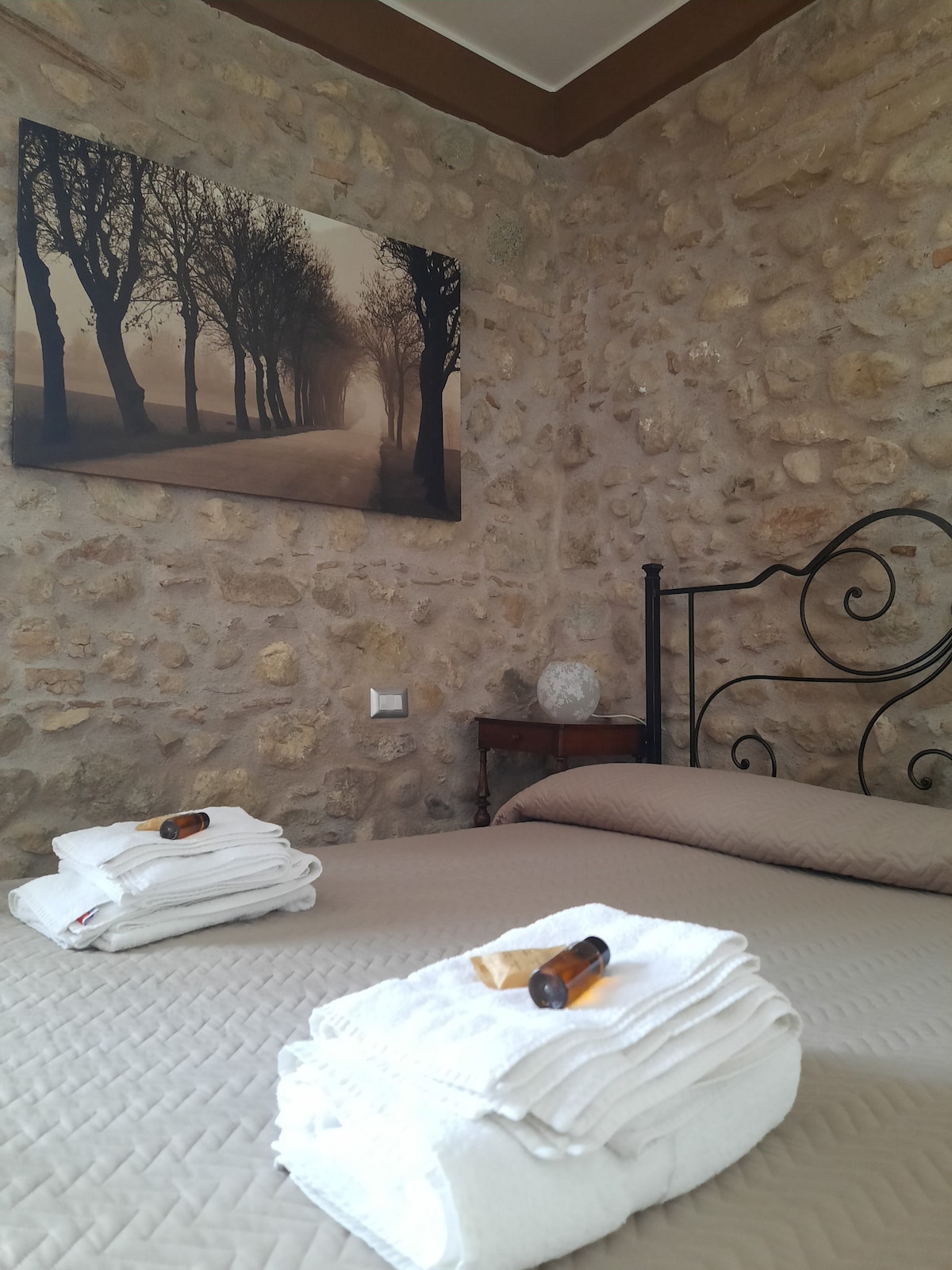 A spacious bed is dressed with neatly arranged white towels. The textured stone wall offers a rustic charm, while a serene artwork of a tree-lined path adds a classic touch to the room. Natural light gently illuminates the space.